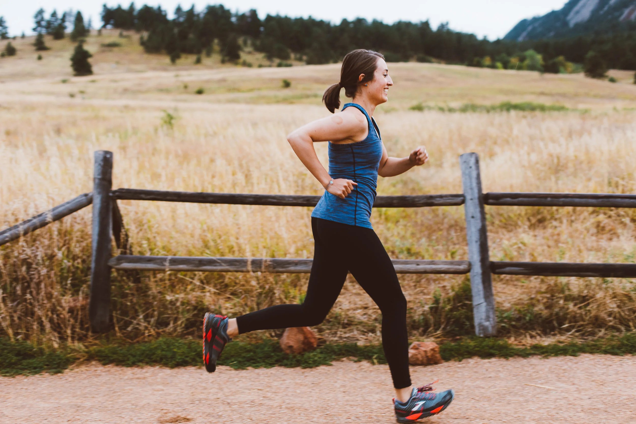 Woman smiling while running along a gravel trail past a wooden fence and grassy field with mountains in the background