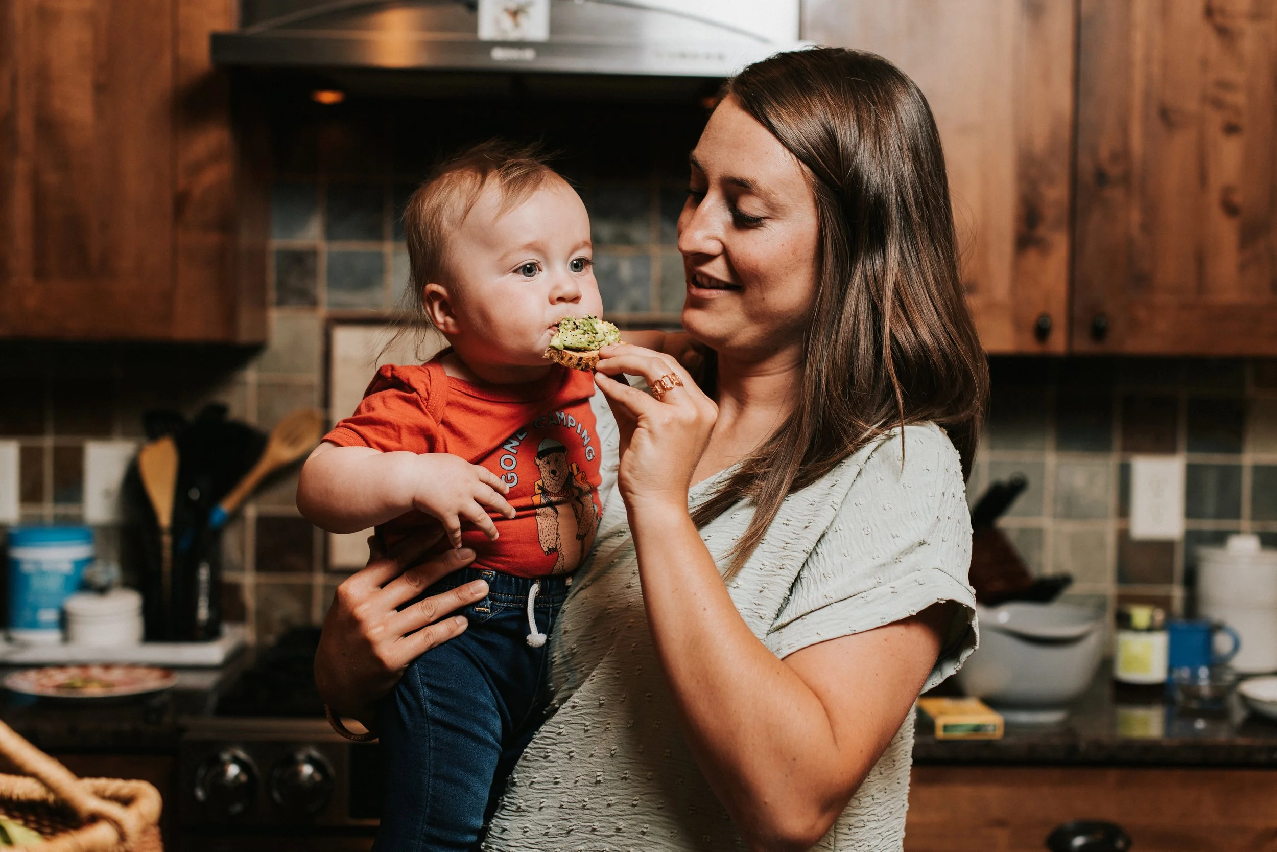 Woman in a kitchen feeding a baby a bite of avocado toast
