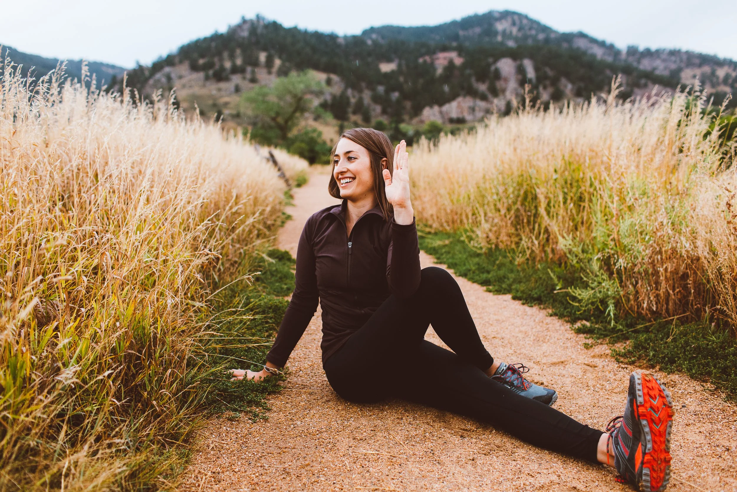 Woman smiling and waving while sitting on a dirt trail through tall grass, mountains in background