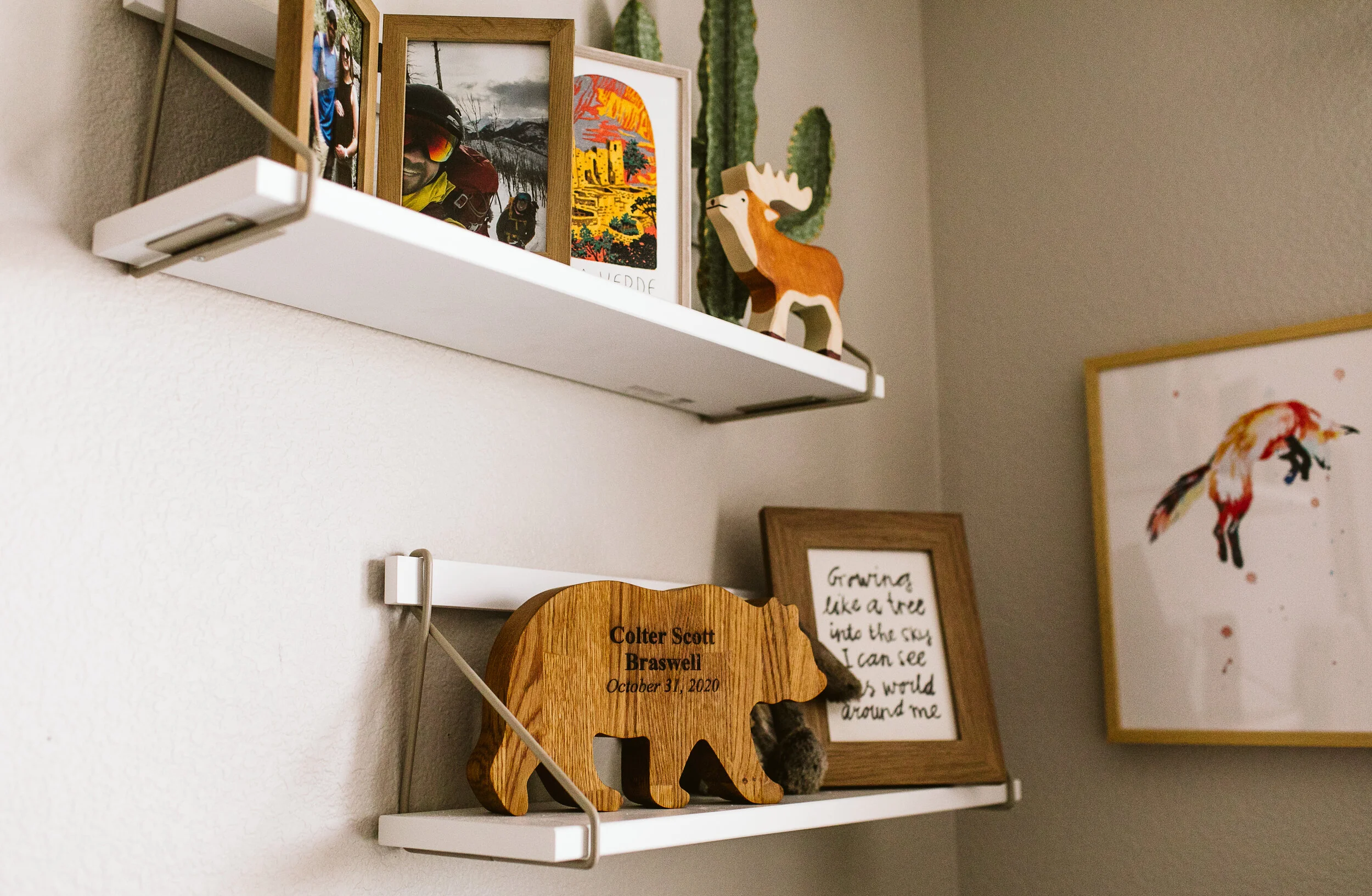 Shelves with framed photos, a wooden moose figure, and a bear-shaped wooden plaque engraved "Colter Scott Braswell, October 31, 2020" beside a framed handwritten quote