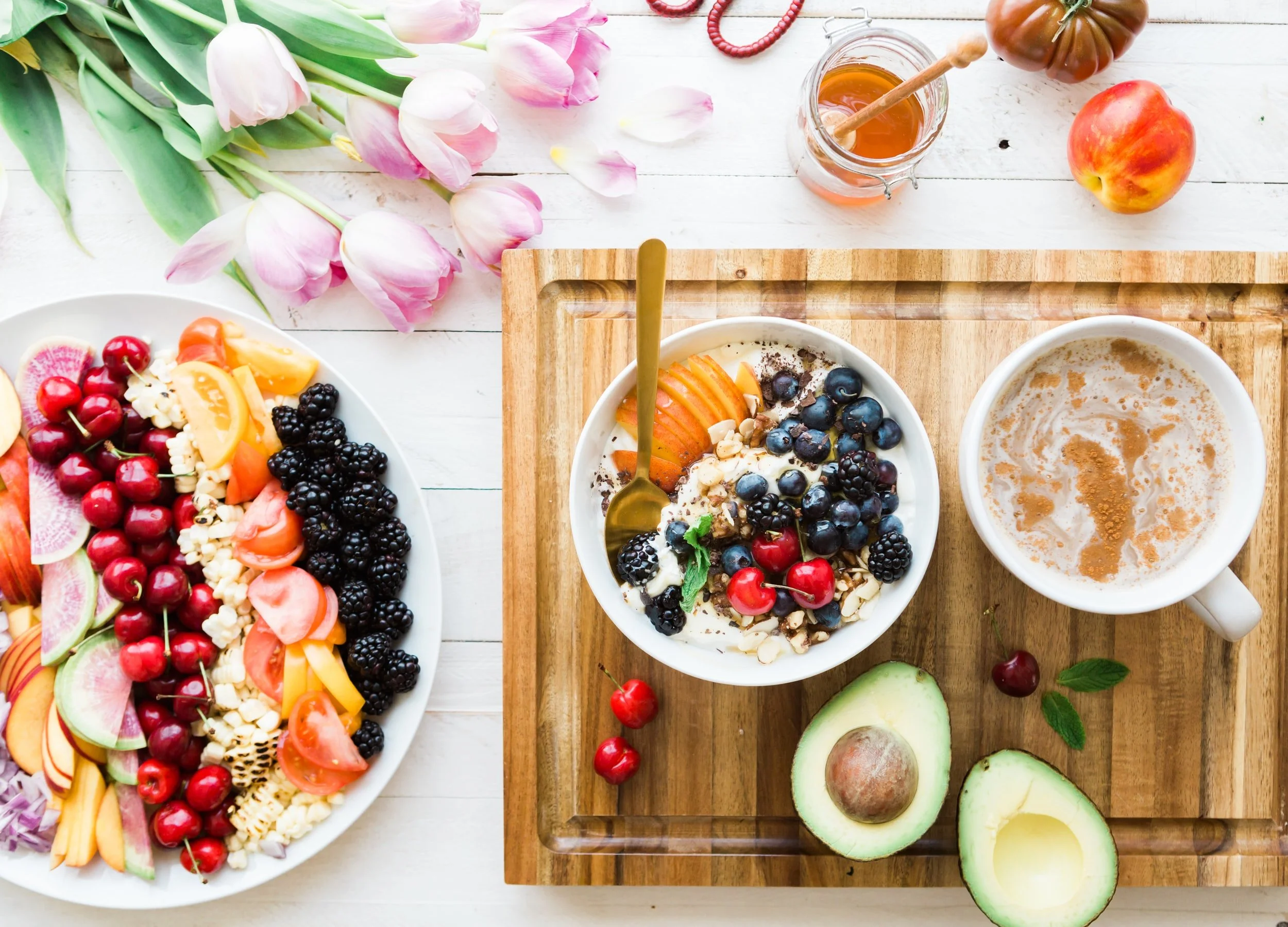 Overhead view of a breakfast spread with a yogurt bowl topped with berries and peach, sliced avocado, a latte, honey jar, fruit and vegetable platter, and pink tulips