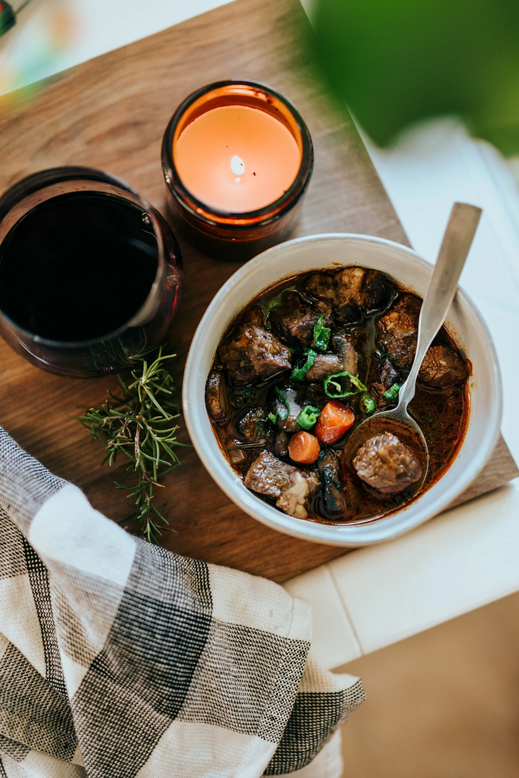 Bowl of beef stew with carrots and mushrooms, served beside red wine, a lit candle, and fresh rosemary