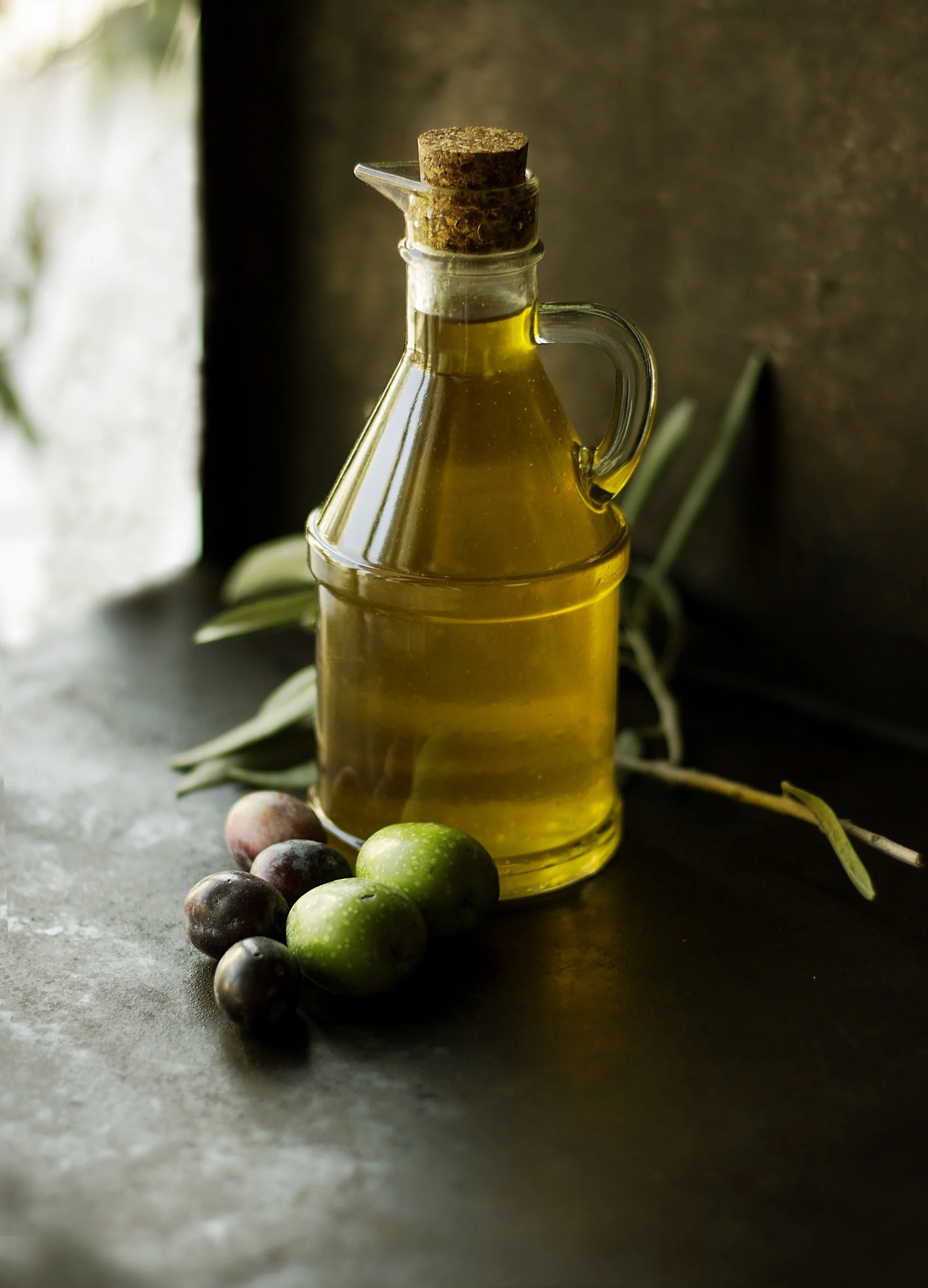 Corked glass bottle of olive oil beside fresh green and black olives on a dark surface