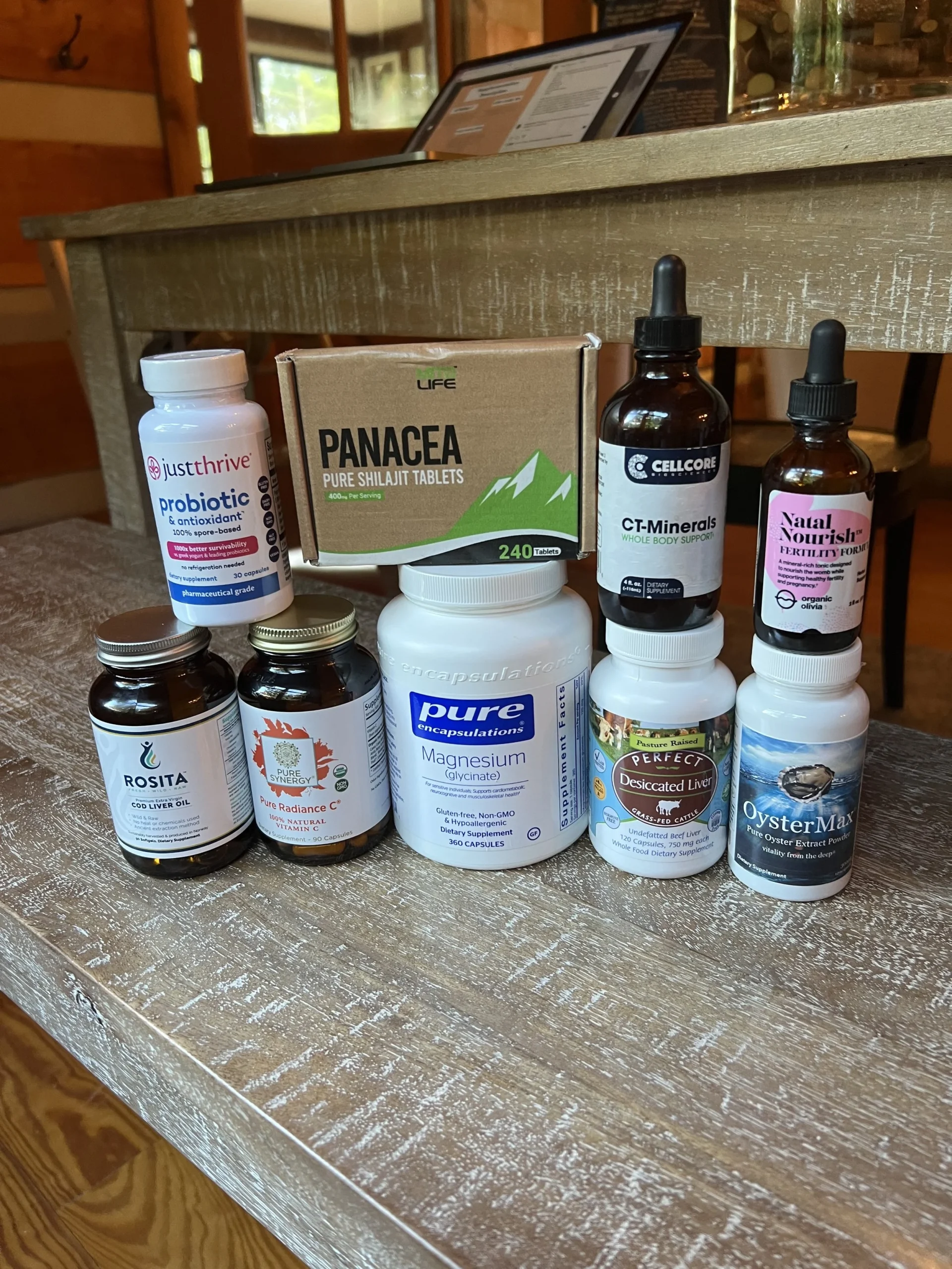 Assortment of supplement bottles including Just Thrive probiotic, Panacea shilajit tablets, Pure Encapsulations magnesium glycinate, CellCore CT-Minerals, Natal Nourish fertility formula, Rosita cod liver oil, Pure Synergy vitamin C, Perfect Desiccated Liver, and OysterMax oyster extract, arranged on a wooden table