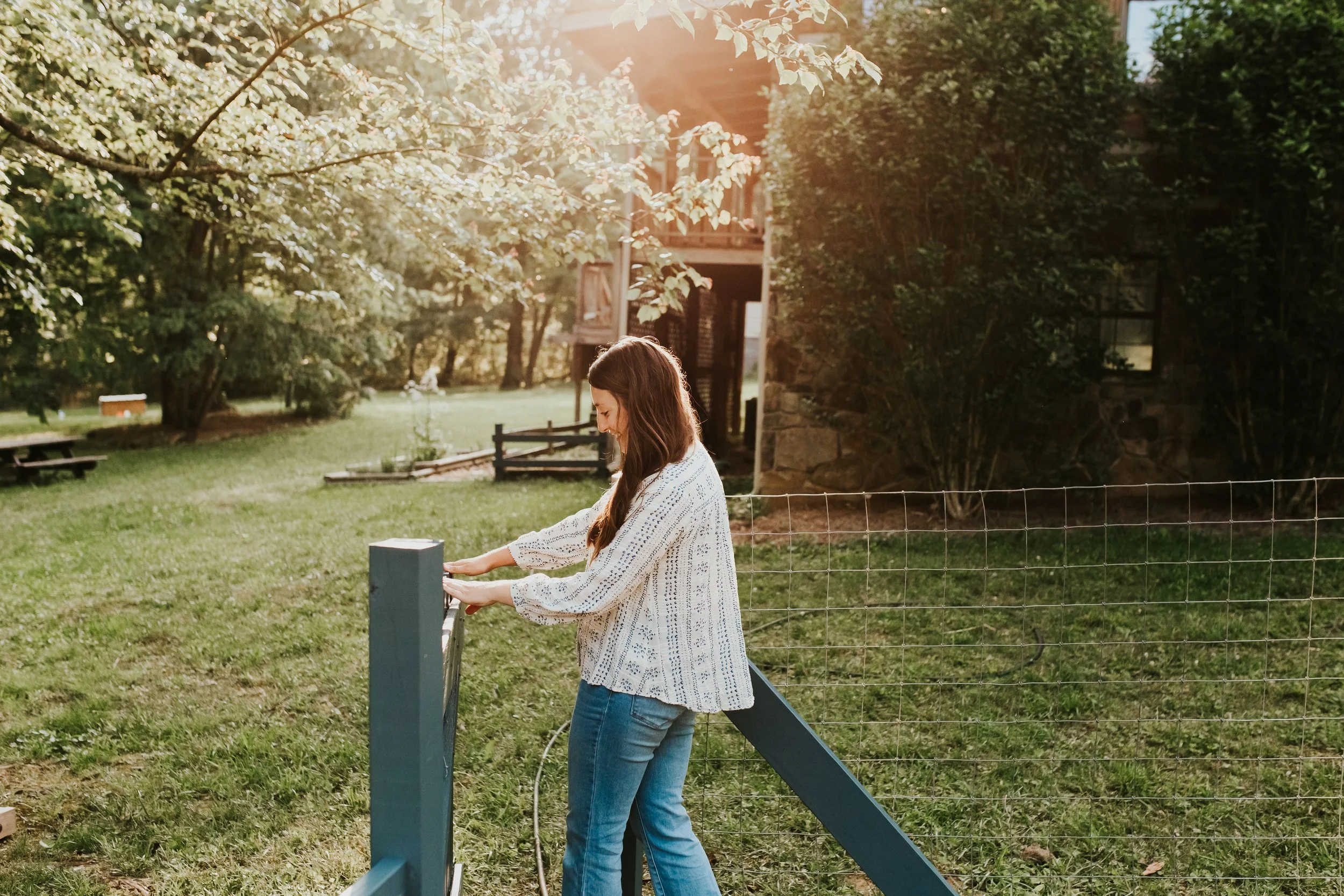 Woman closing a wire garden fence gate in a sunlit backyard near a stone building
