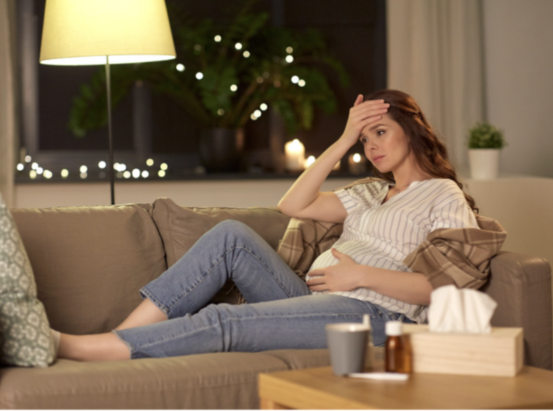 Pregnant woman lying on a couch holding her forehead, looking unwell, with tissues and medicine nearby