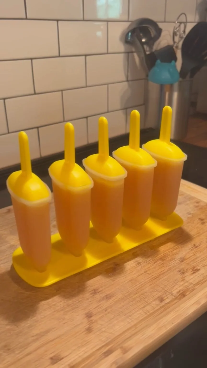 Five orange-colored popsicles freezing in a yellow plastic mold on a wooden kitchen counter