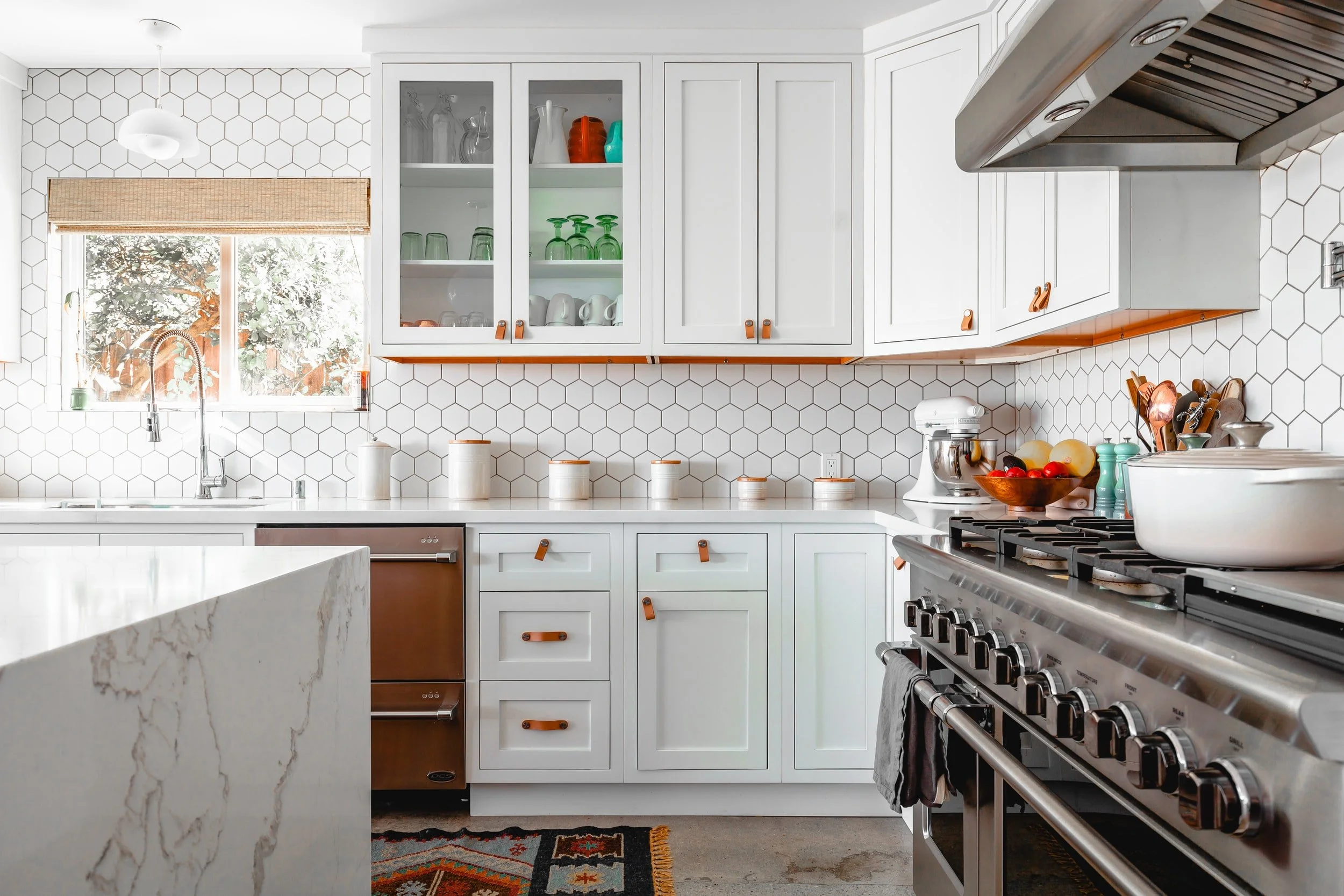 White kitchen with hexagon tile backsplash, leather cabinet pulls, marble island, and stainless steel range with copper appliances
