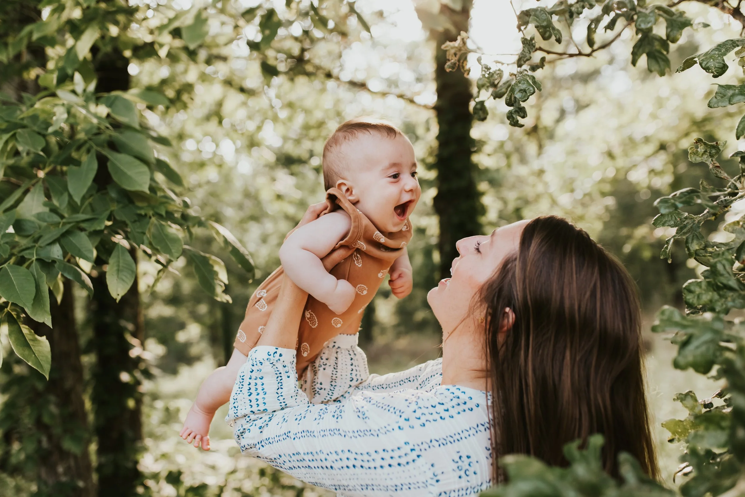Mother lifting a laughing baby into the air outdoors amid green trees
