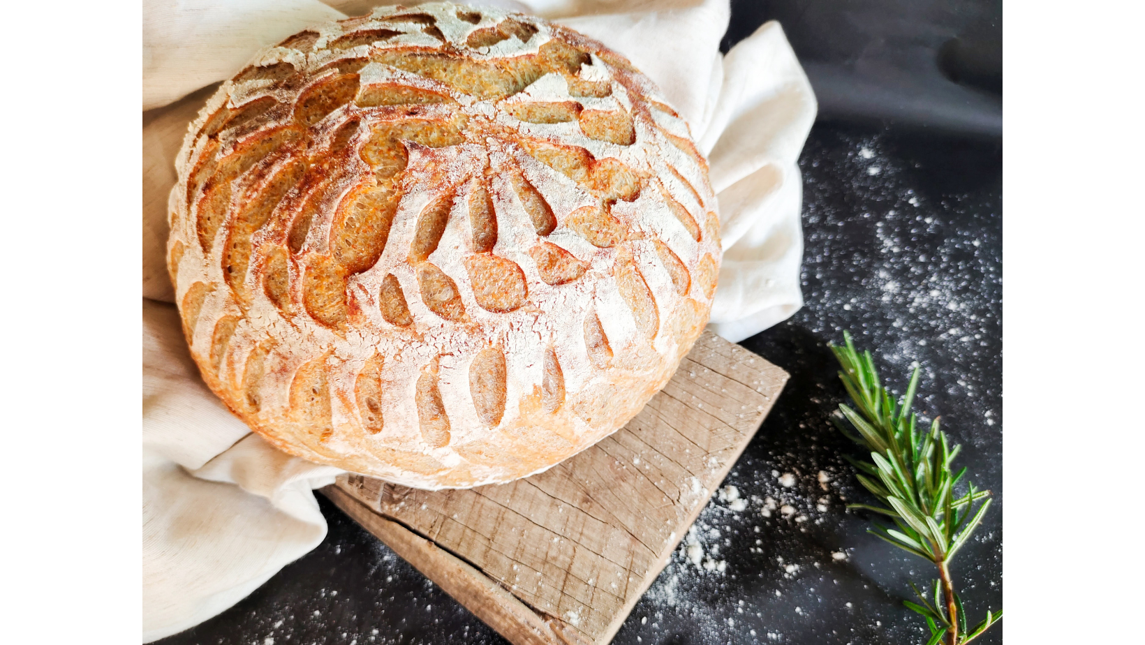Round sourdough loaf with a decorative leaf-pattern scored crust, resting on a wooden board with a rosemary sprig and scattered flour nearby