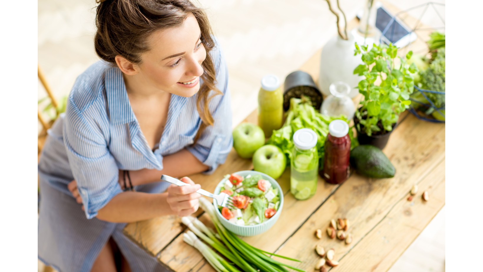 Woman smiling while eating a salad at a table surrounded by fresh vegetables, fruit, herbs, and juices