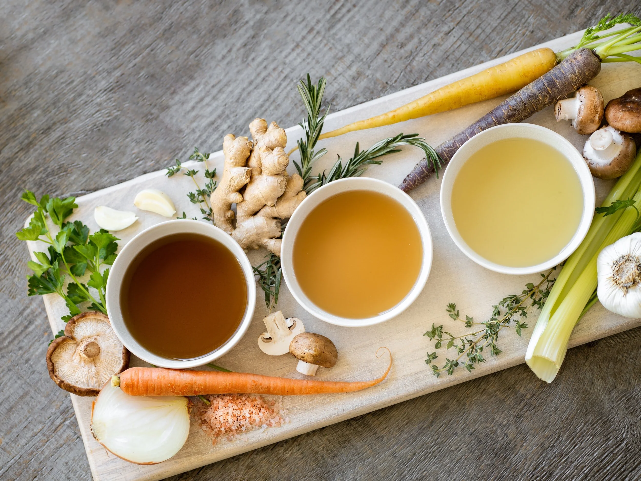 Three bowls of broth in varying shades, surrounded by ginger, carrots, mushrooms, celery, garlic, onion, and herbs