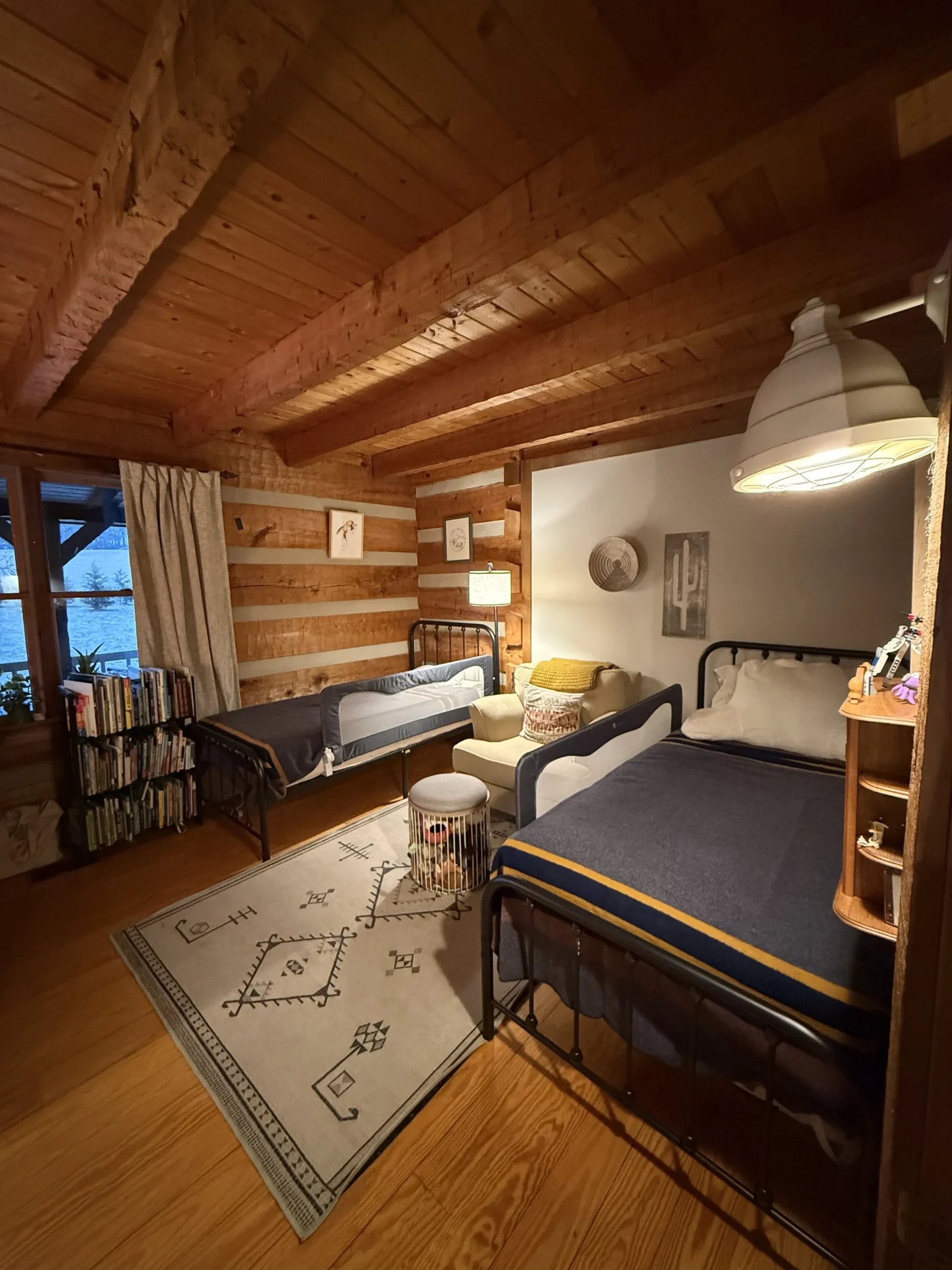 Log cabin bedroom with two twin metal-frame beds, a patterned rug, bookshelf, armchair, and wooden beamed ceiling