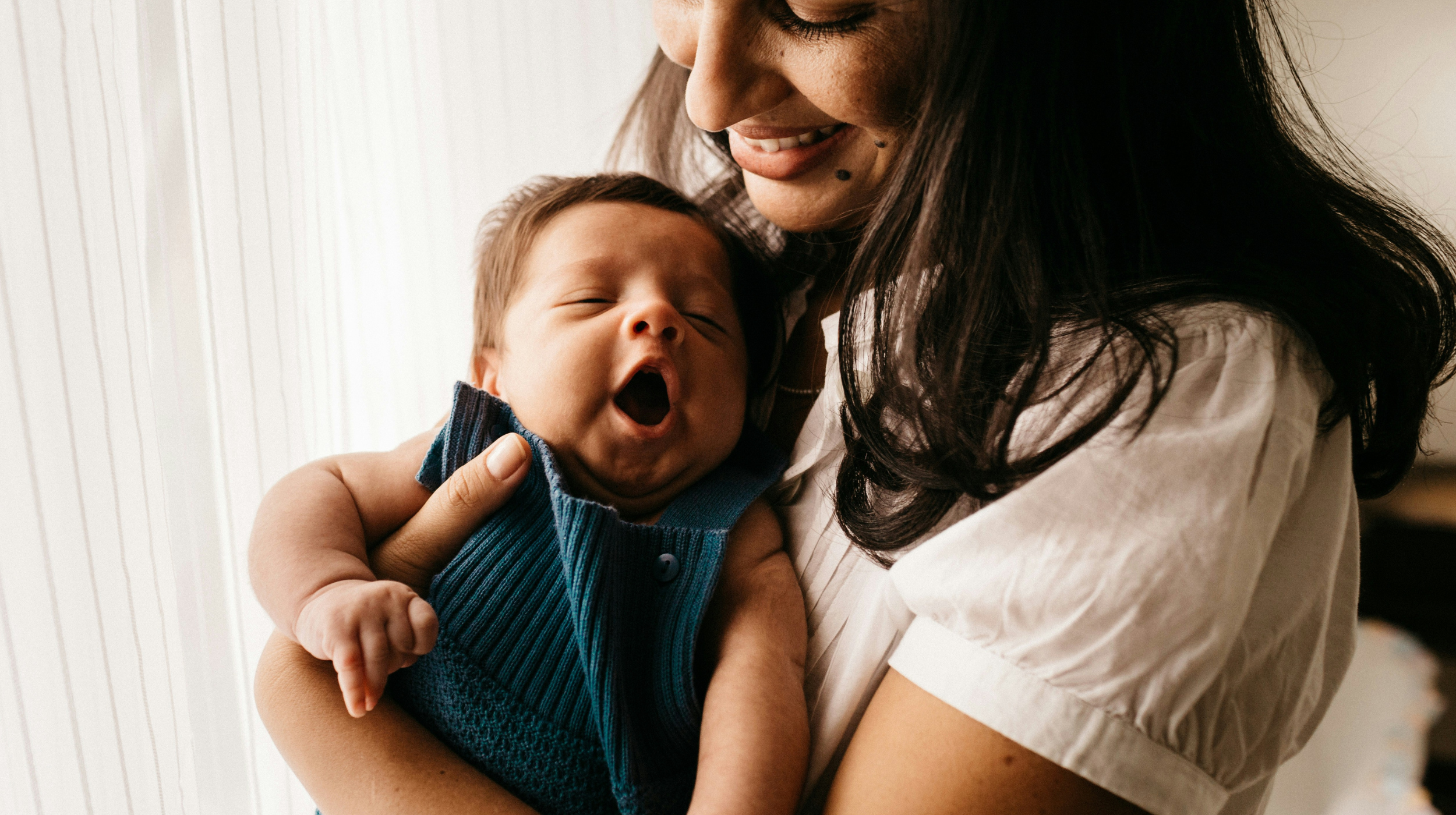Smiling woman holds a yawning newborn in a blue knit outfit near a sunlit window