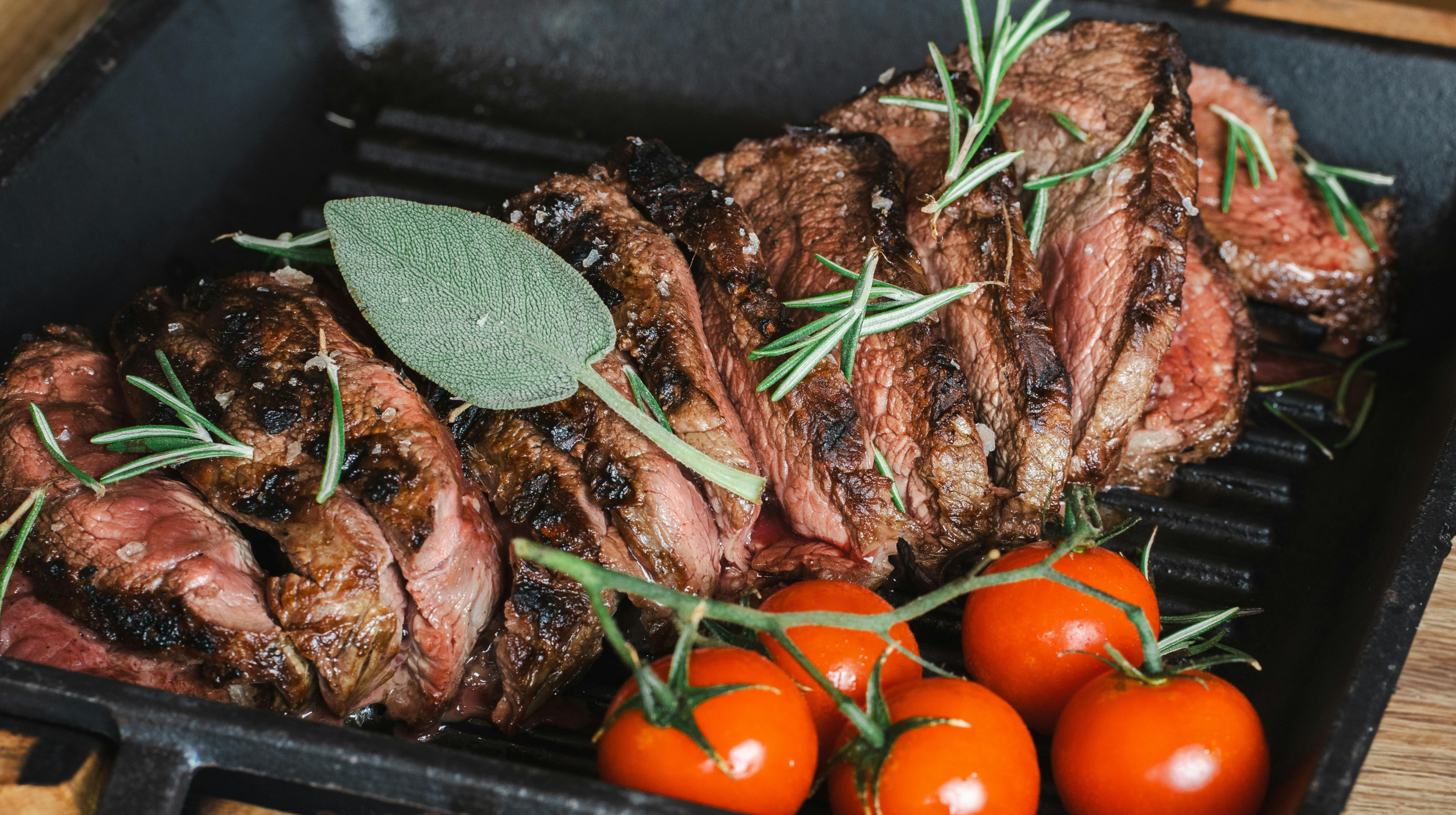 Sliced grilled steak with rosemary and sage on a cast-iron grill pan, alongside a bunch of vine tomatoes