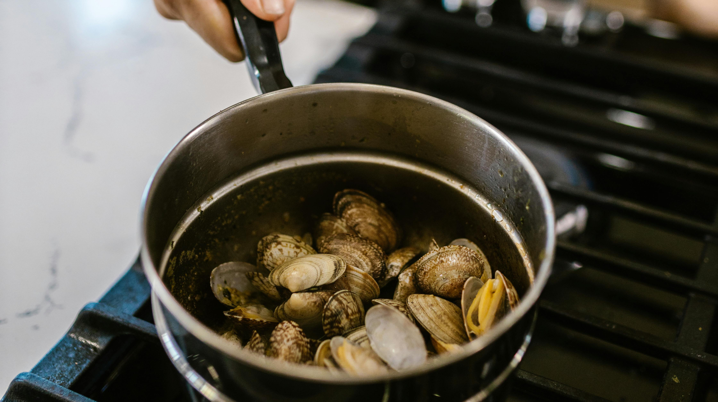 Pot of open clams cooking with pasta on a stovetop, held with tongs