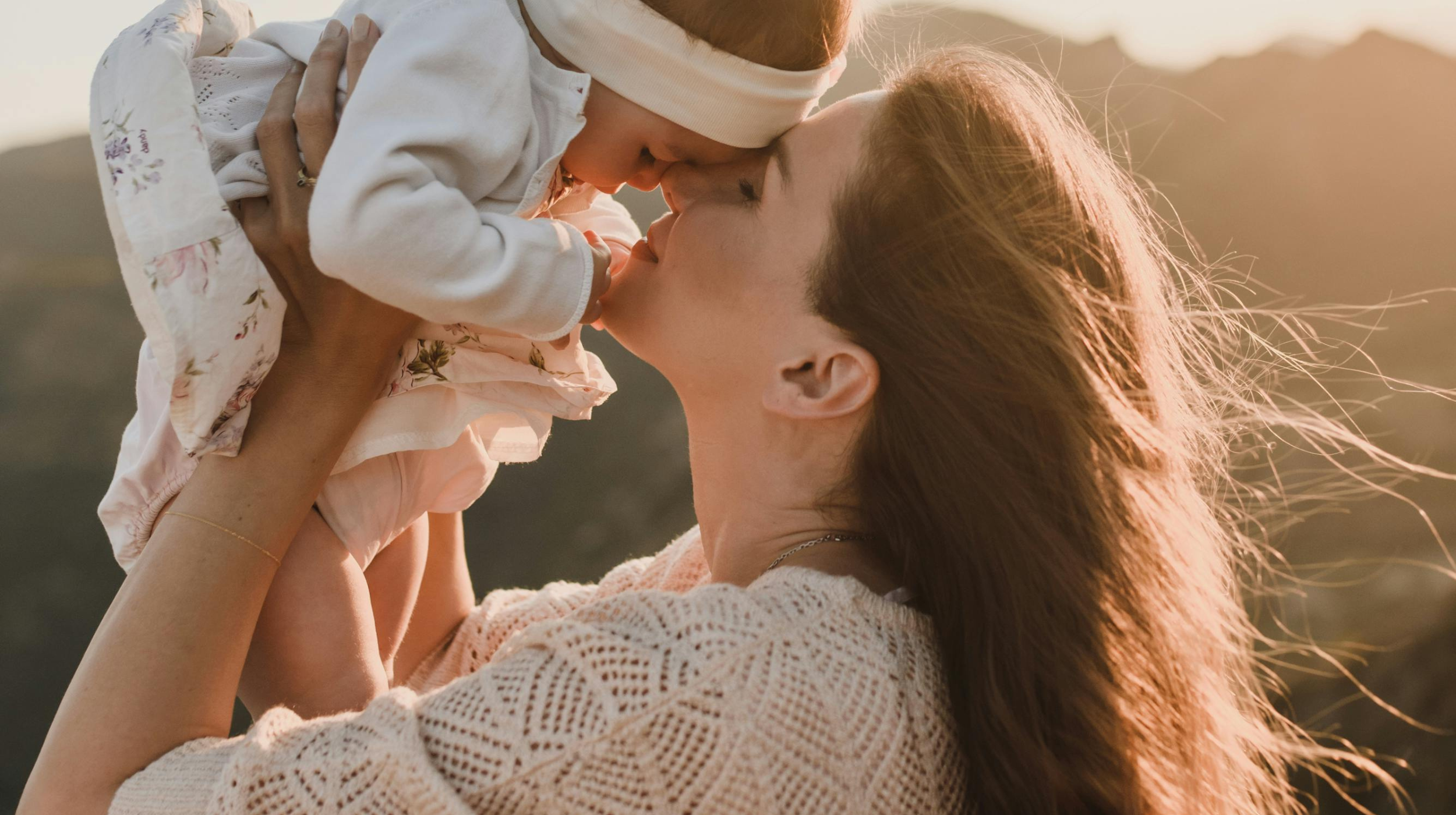 Mother lifting and kissing her baby's nose outdoors at golden hour, mountains in background