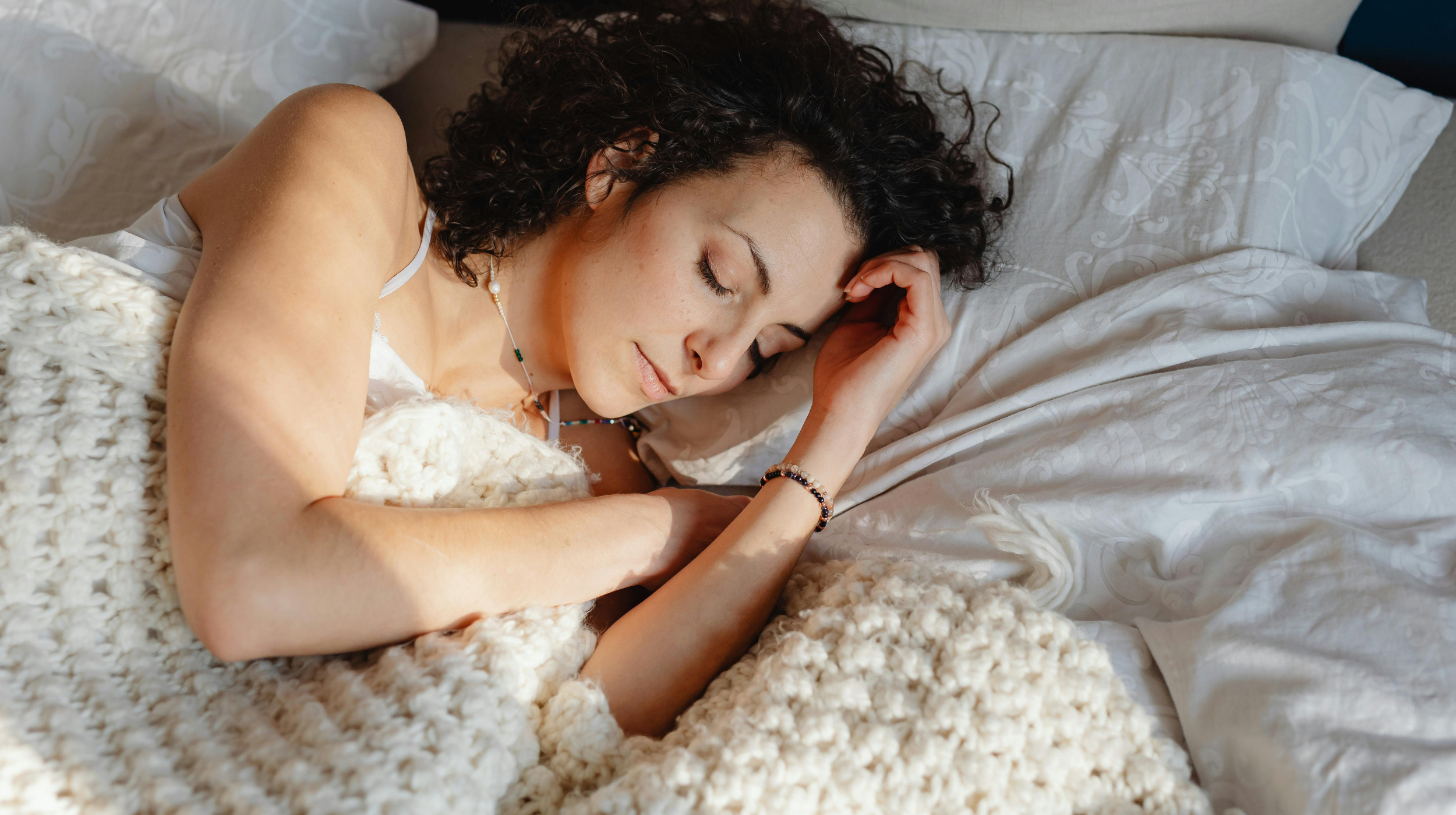 Woman with curly hair sleeping peacefully in bed, sunlight falling across her face and arms