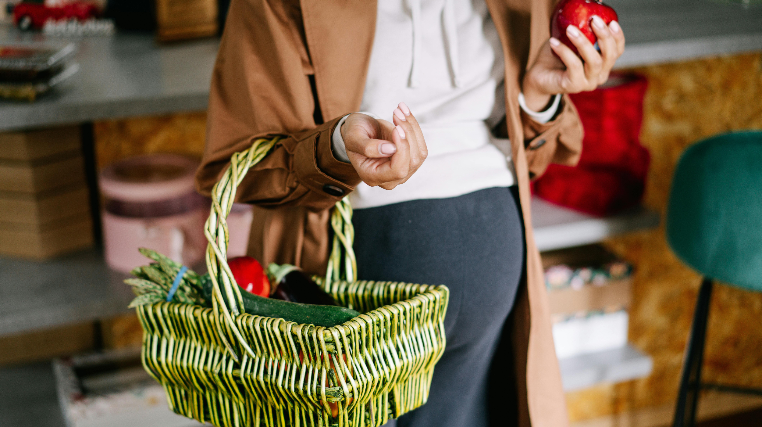 Person carrying a woven basket of vegetables, holding an apple in the other hand