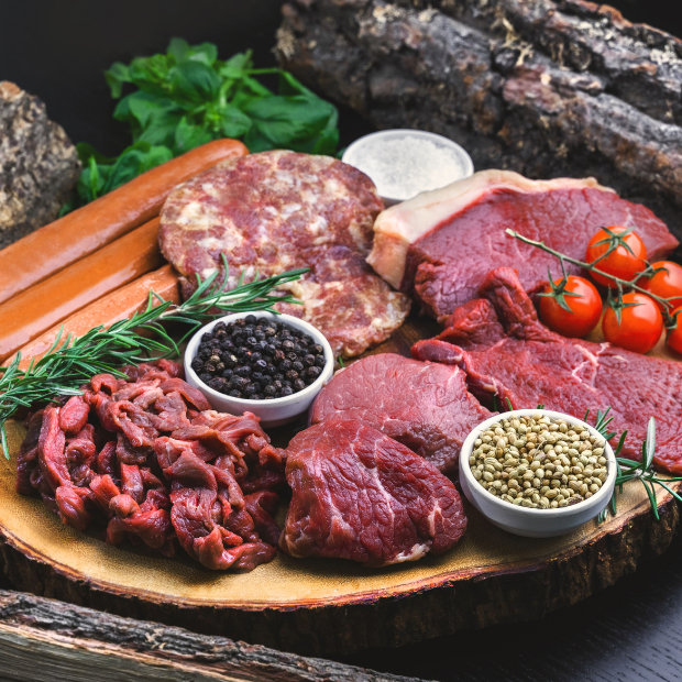 Assortment of raw meats including steak, sausages, and sliced beef with peppercorns, coriander seeds, cherry tomatoes, and herbs on a wooden board