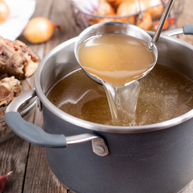 Ladle pouring golden bone broth from a large pot, with roasted bones and onions nearby