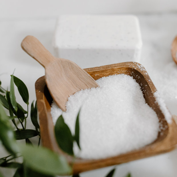 Wooden bowl of coarse white salt or bath crystals with a wooden scoop, beside greenery and a soap bar