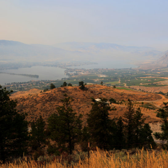 Hazy valley view of a lake surrounded by farmland, hills, and mountains under smoky skies