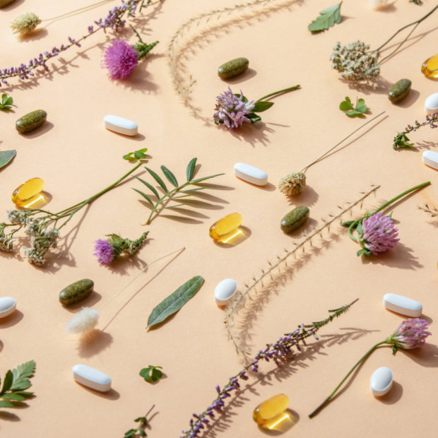 Assorted vitamin capsules, softgels, and tablets arranged among dried flowers and herbs on a peach background