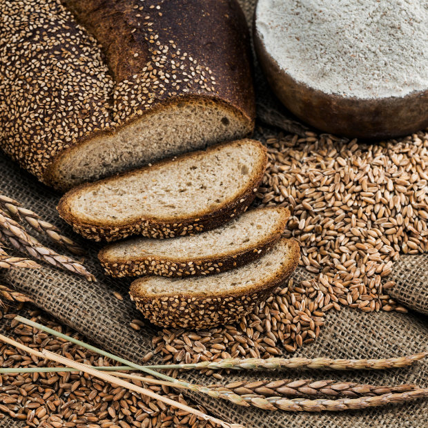 Sliced sesame-crusted bread loaf surrounded by wheat grains, stalks, and a bowl of flour