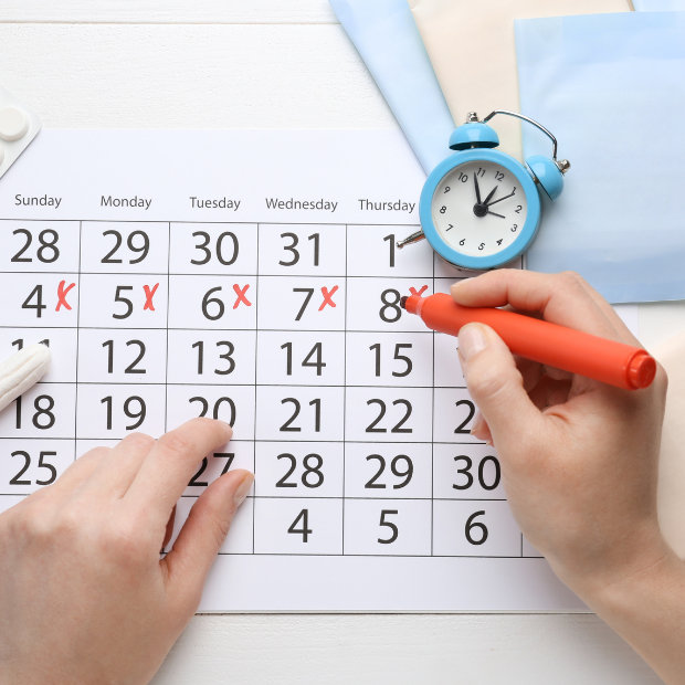 Hands marking off days on a calendar with a red marker, tracking a menstrual cycle, with alarm clock nearby