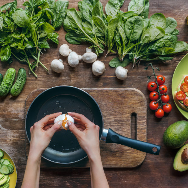 Hands cracking an egg into a pan, surrounded by spinach, mushrooms, cucumbers, cherry tomatoes, and avocados