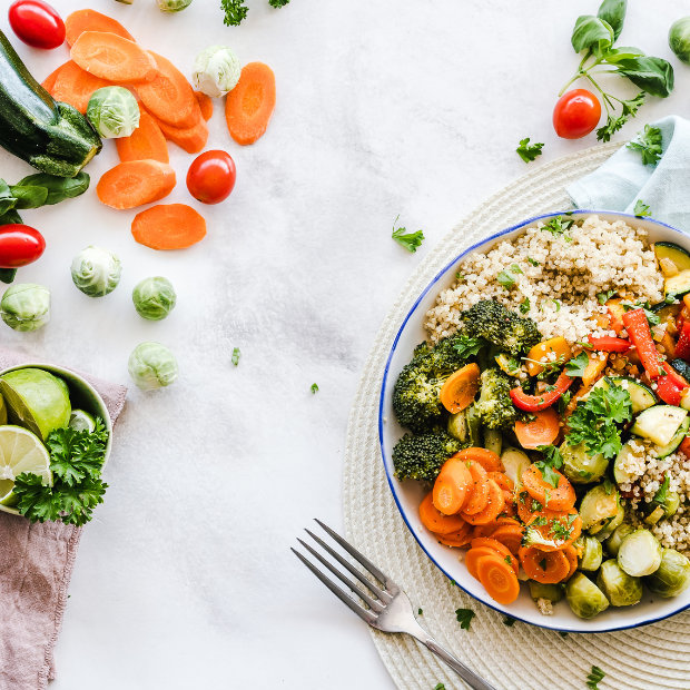Bowl of quinoa with roasted broccoli, carrots, brussels sprouts, and peppers, surrounded by fresh vegetables
