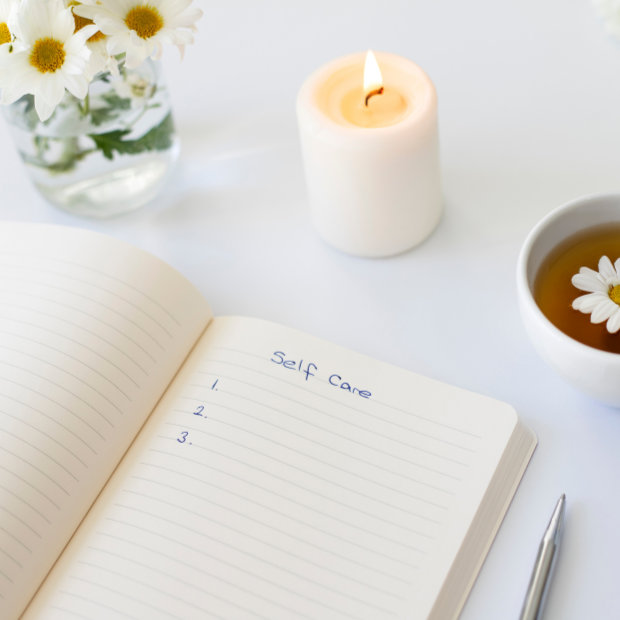 Open notebook with a "Self Care" numbered list, beside a lit candle, daisies, and a cup of tea