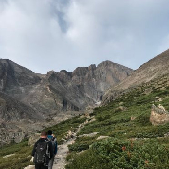 Two hikers with backpacks walk a rocky trail toward jagged mountain peaks under cloudy sky