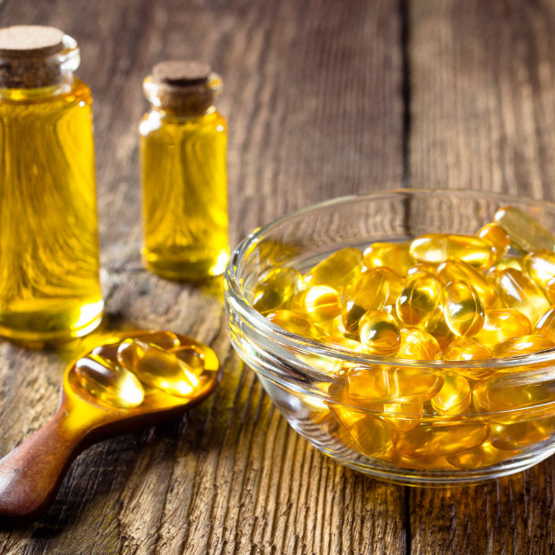 Bowl and spoon of golden fish oil capsules beside bottles of oil on wood table