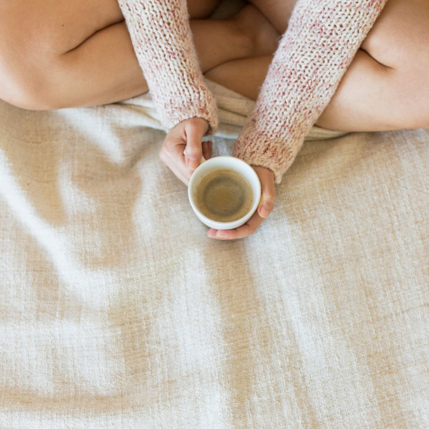 Person in knit sleeve warmers sitting cross-legged, holding a small cup of coffee on a blanket
