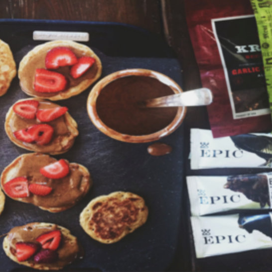 Mini pancakes topped with strawberries and nut butter next to a bowl of chocolate sauce and Epic protein bar packages
